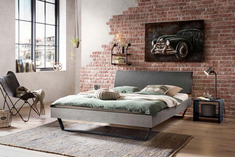 Ein modernes Schlafzimmer mit einer Akzentwand aus Backstein, einem großen Fenster, einem minimalistischen Bett mit grüner Bettwäsche, einem Gemälde mit einem Oldtimer, einem Stuhl, einem Beistelltisch, einer Lampe und einem gewebten Teppich auf hellem Holzboden.