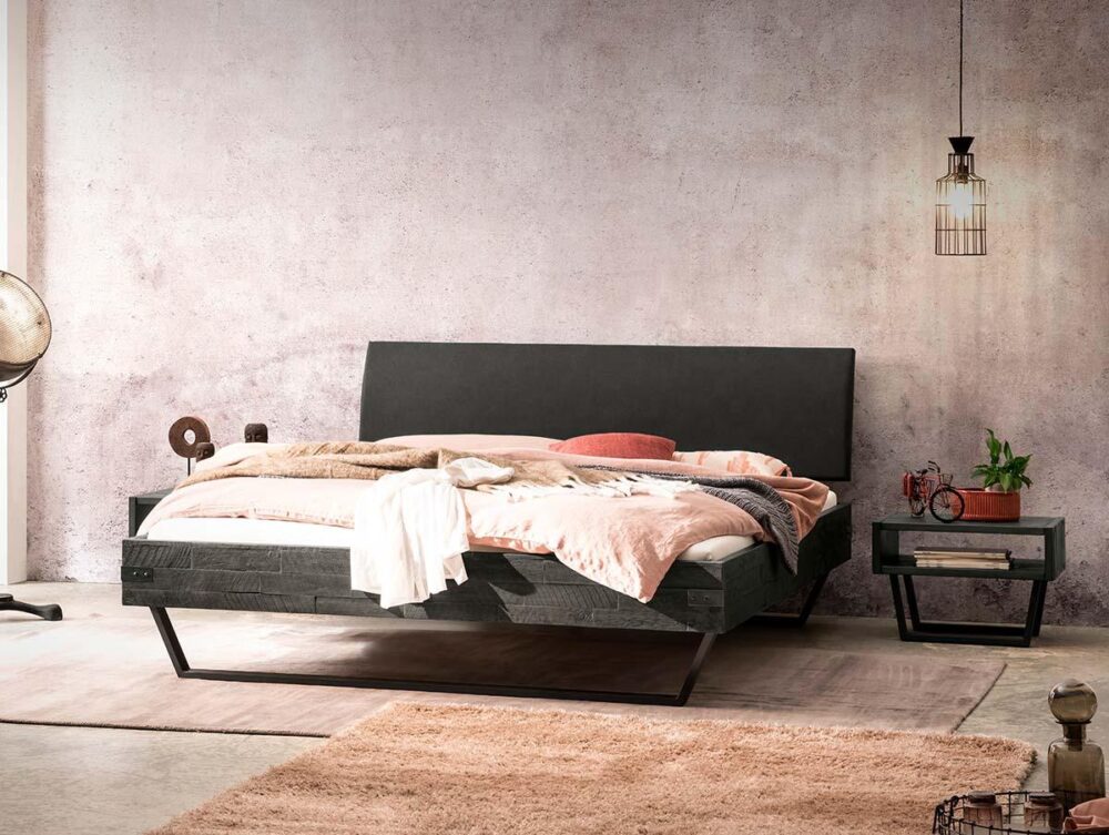 Ein modernes Schlafzimmer mit einem Bettgestell aus dunklem Holz, neutraler Bettwäsche, einer rosa Decke, einem Beistelltisch mit einer Pflanze, einer von der Decke hängenden Lampe und einer strukturierten hellrosa Wand. Das Licht fällt von links ein.