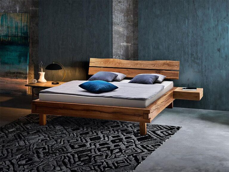 Ein modernes Schlafzimmer mit einem Holzbettgestell, einer grauen Matratze und blauen und grauen Kissen. Das Bett steht auf einem schwarz-weiß gemusterten Teppich neben einem Nachttisch mit einer Lampe, Büchern und einer Vase vor blau-grauen, strukturierten Wänden.