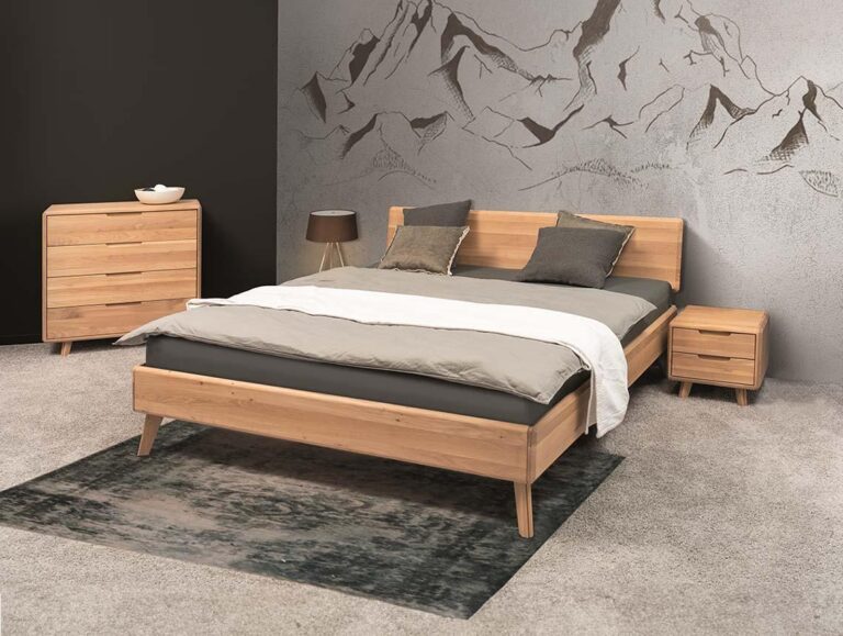 Modernes Schlafzimmer mit einem hölzernen Bettgestell, zweifarbiger Bettwäsche, drei Kissen, einem Nachttisch, einer Kommode mit einer Schale darauf, einem gemusterten Teppich und einer Wandmalerei mit Bergen im Hintergrund.