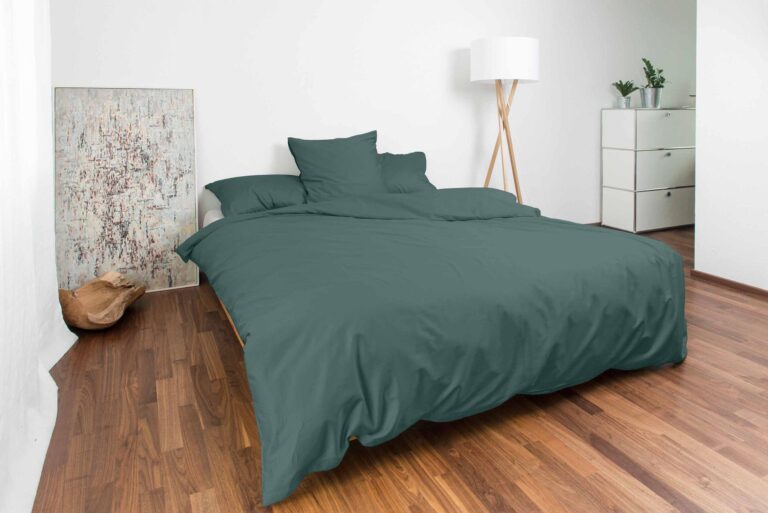 Ein modernes Schlafzimmer mit einem Doppelbett, bezogen mit der tealfarbenen Bettwäsche Nobilium Oni, zwei Kissen und einem Kissen. Der Raum hat auch einen Holzboden, eine weiße Stehlampe, eine Kommode mit Pflanzen und ein Gemälde an der Wand.