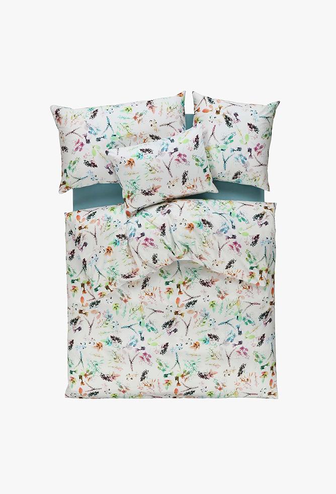 Das Bettwäsche-Set Tamara-R Selection Lissy ist auf einem Bett mit weißer Bettdecke und Kissen mit lebhaftem Blumen- und Blättermuster in Grün, Orange, Violett und Blau abgebildet, von oben gesehen auf einem schlichten weißen Hintergrund.