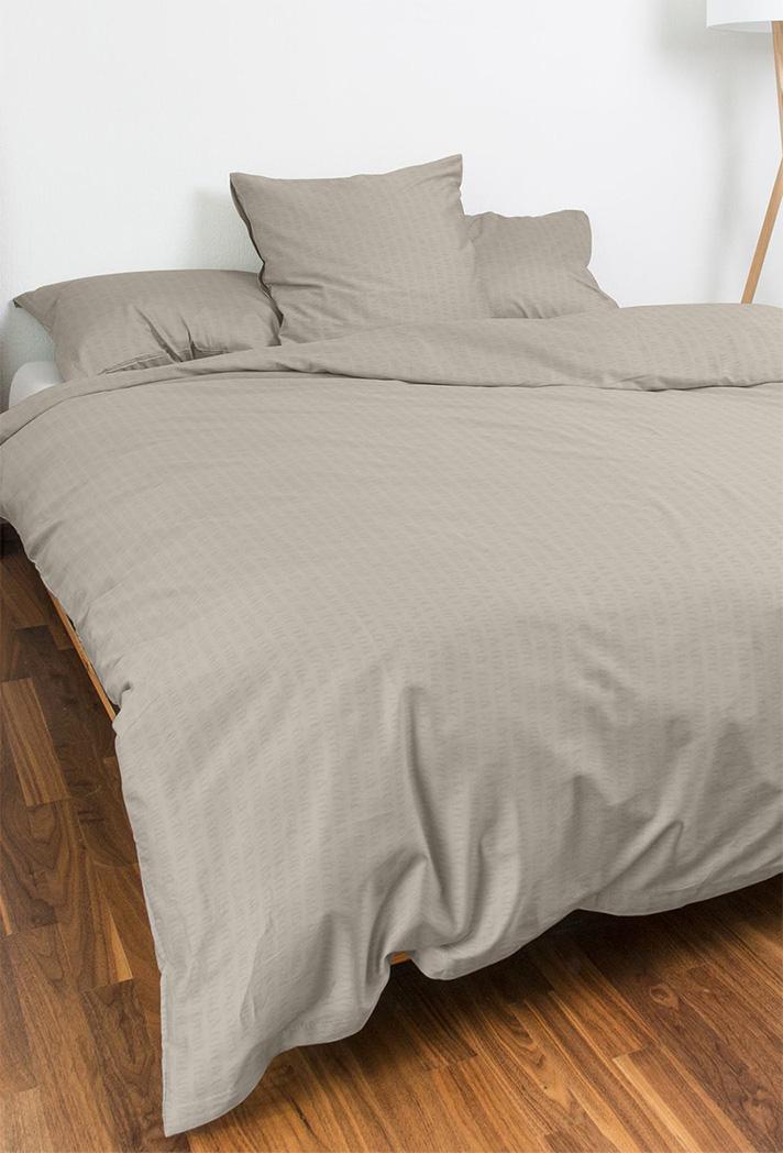 Ein ordentlich gemachtes Bett mit Bettwäsche Night Fashion Edi in Hellbeige, mit Bettbezug und Kissen, steht auf einem Holzboden. Im Hintergrund steht eine weiße Lampe mit einem Holzfuß.