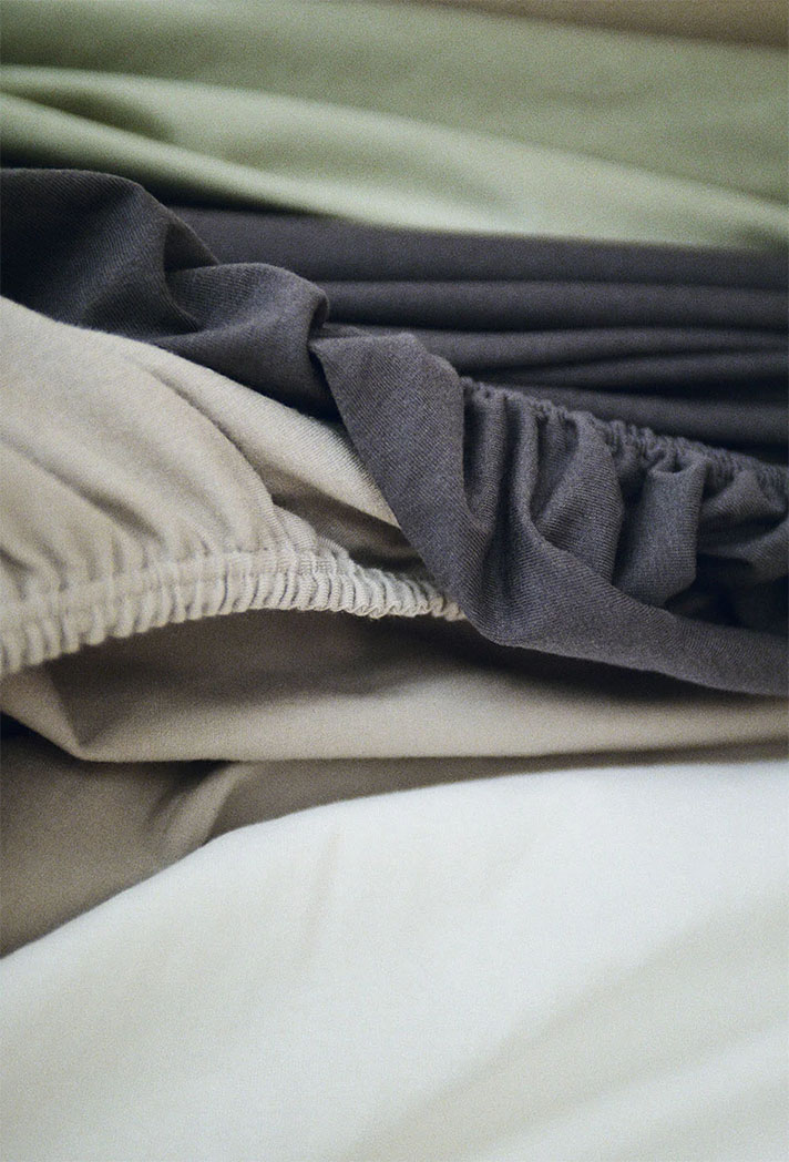 Eine Nahaufnahme mehrerer geschichteter, zerknitterter Bettlaken und Spannbettlaken in neutralen Tönen wie Beige, Grau und Off-White, die die Textur des Stoffes und die elastischen Kanten zeigen.