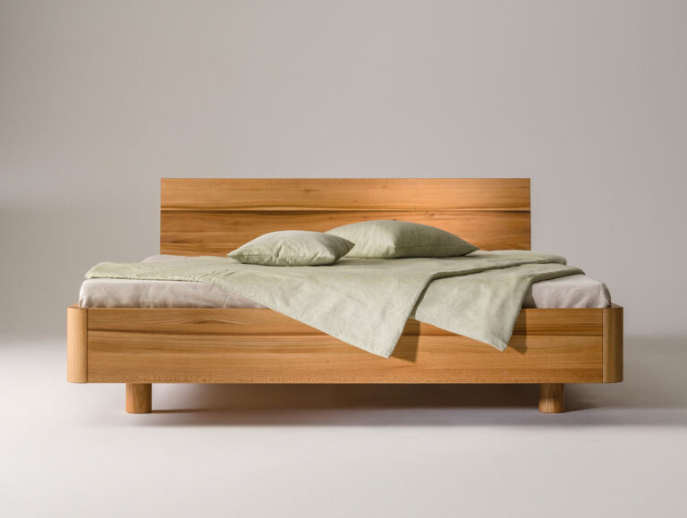 Ein modernes Holzbett mit einem einfachen Kopfteil, hellgrüner Bettwäsche und zwei passenden Kissen vor einem schlichten weißen Hintergrund.