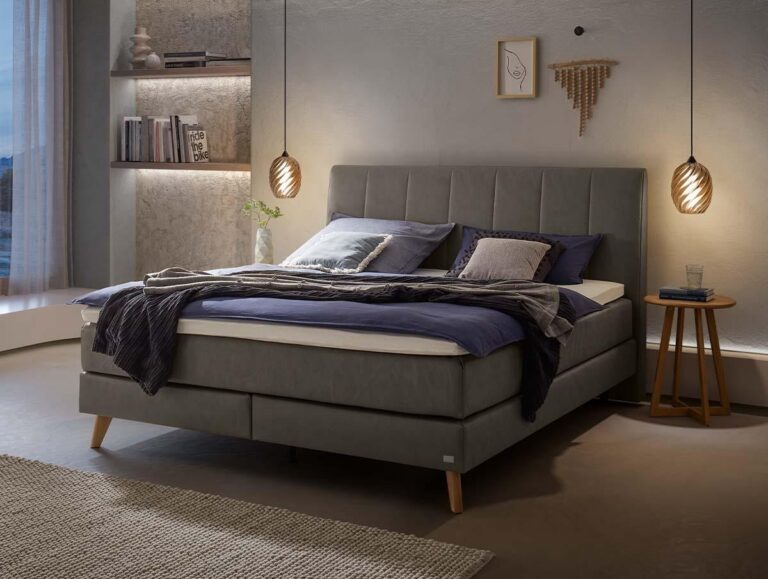 Ein modernes Schlafzimmer verfügt über ein ordentlich gemachtes graues Bett mit blauer und grauer Bettwäsche, zwei Pendelleuchten, einen hölzernen Nachttisch mit einem Wasserglas, Wandkunst und Regale mit Büchern und Dekoration.