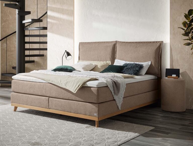 Ein modernes Schlafzimmer mit einem beigefarbenen gepolsterten Bett, das mit Kissen und einer Decke ausgelegt ist. Neben dem Bett steht ein Nachttisch mit einer Lampe und Dekoration, und im Hintergrund ist eine Wendeltreppe zu sehen. In der Ecke steht eine Pflanze.