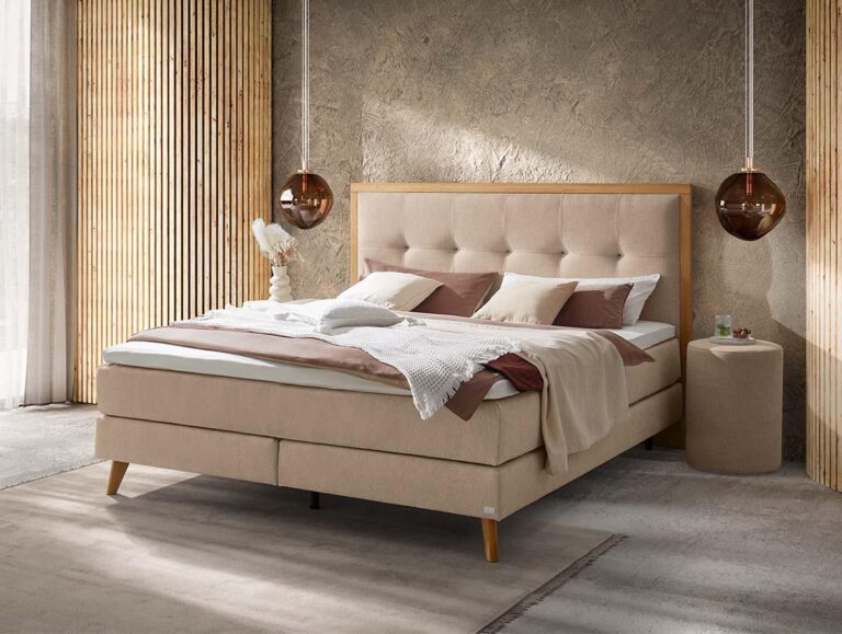 Ein modernes Schlafzimmer mit einem beigen Polsterbett, brauner und weißer Bettwäsche, zwei braunen Hängelampen, einem runden beigen Nachttisch und Holzlatten an der Wand. Natürliches Licht fällt durch ein Fenster mit durchsichtigen Vorhängen ein.