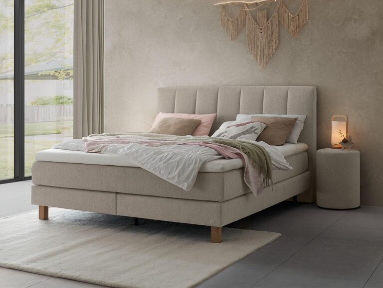 Ein modernes, beigefarbenes Polsterbett mit weißer und rosa Bettwäsche steht auf einem hellen Teppich in einem minimalistischen Schlafzimmer. Eine sanfte Lampe steht auf einem kleinen runden Tisch neben dem Bett, mit großen Fenstern und einem Makramee-Wandbehang im Hintergrund.