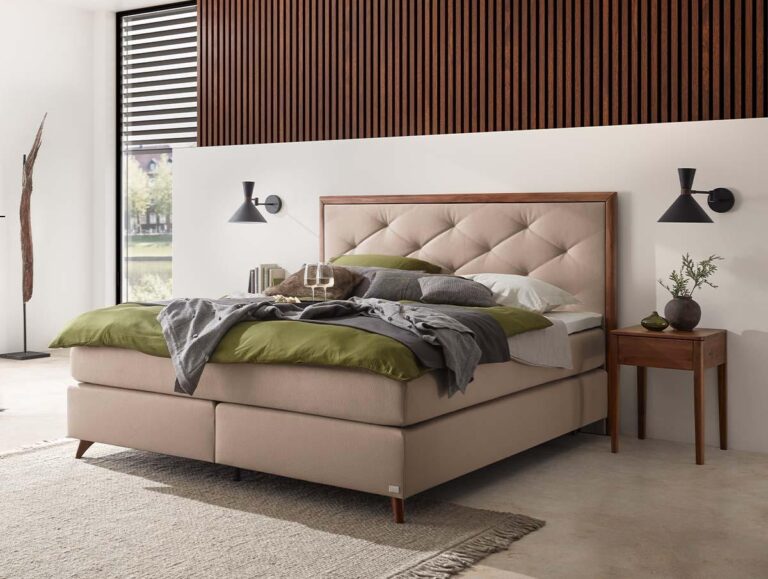 Ein modernes Schlafzimmer mit einem beigen Polsterbett, grüner und grauer Bettwäsche, einem getufteten Kopfteil, einem hölzernen Nachttisch mit einer Vase und schwarzen Wandleuchten. Die Wand hinter dem Bett ist mit einer vertikalen Holzvertäfelung verziert.