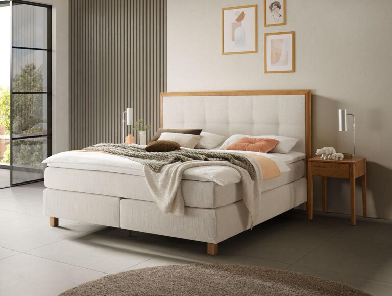 Ein modernes Schlafzimmer mit einem großen Polsterbett, beiger Bettwäsche, einem hölzernen Nachttisch mit einer Lampe, gerahmter Kunst an der Wand und einem runden braunen Teppich auf einem gefliesten Boden. Das Sonnenlicht scheint durch ein großes Fenster.
