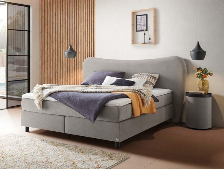 Ein modernes Schlafzimmer mit einem großen grauen Bett, mehreren Kissen und Decken, zwei schwarzen Pendelleuchten, einem runden Nachttisch mit einer Blumenvase und einem Teppich auf einem hellen Boden. Hölzerne und weiße Wände sorgen für eine gemütliche Atmosphäre.