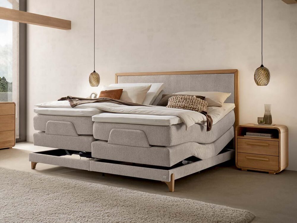 Ein modernes Schlafzimmer mit einem hellen, verstellbaren Polsterbett, beiger Bettwäsche und zwei Pendelleuchten, die über hölzernen Nachttischen hängen. Das Sonnenlicht fällt durch ein großes Fenster und schafft eine warme, gemütliche Atmosphäre.