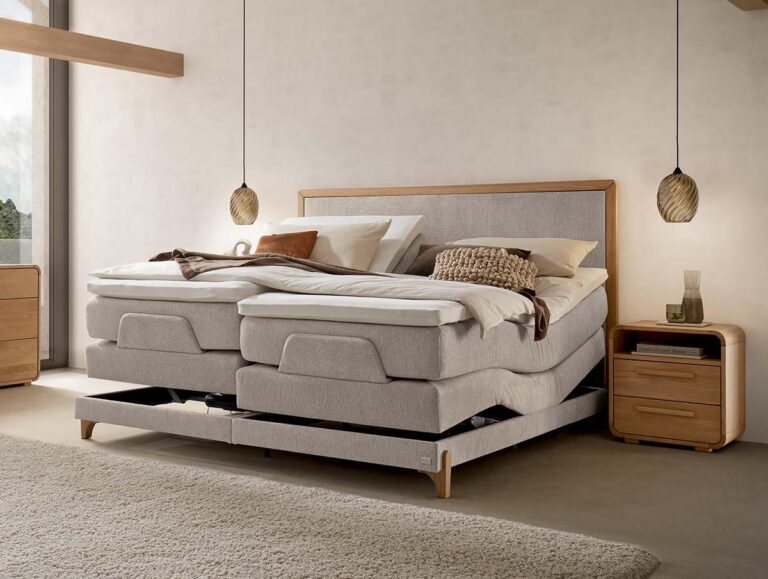 Ein modernes Schlafzimmer mit einem hellen, verstellbaren Polsterbett, beiger Bettwäsche und zwei Pendelleuchten, die über hölzernen Nachttischen hängen. Das Sonnenlicht fällt durch ein großes Fenster und schafft eine warme, gemütliche Atmosphäre.