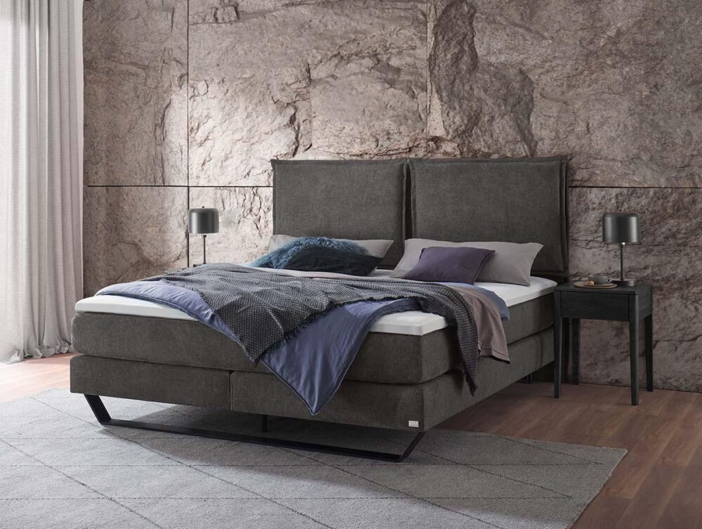 Ein modernes Schlafzimmer mit einem dunkelgrauen gepolsterten Bett, blauer und grauer Bettwäsche, zwei Kissen und zwei schwarzen Nachttischen mit Lampen. Das Zimmer hat eine Akzentwand mit Steinstruktur, helle Vorhänge und einen grauen Teppich auf Holzfußboden.