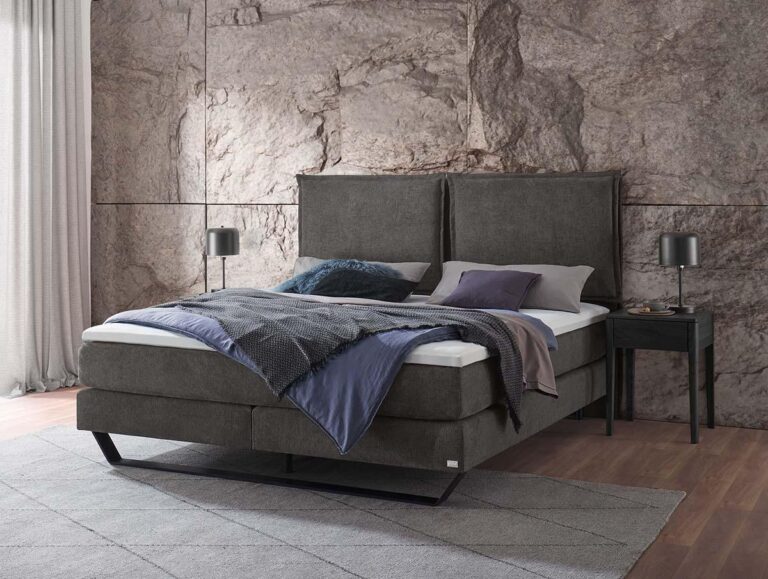 Ein modernes Schlafzimmer mit einem dunkelgrauen gepolsterten Bett, blauer und grauer Bettwäsche, zwei Kissen und zwei schwarzen Nachttischen mit Lampen. Das Zimmer hat eine Akzentwand mit Steinstruktur, helle Vorhänge und einen grauen Teppich auf Holzfußboden.