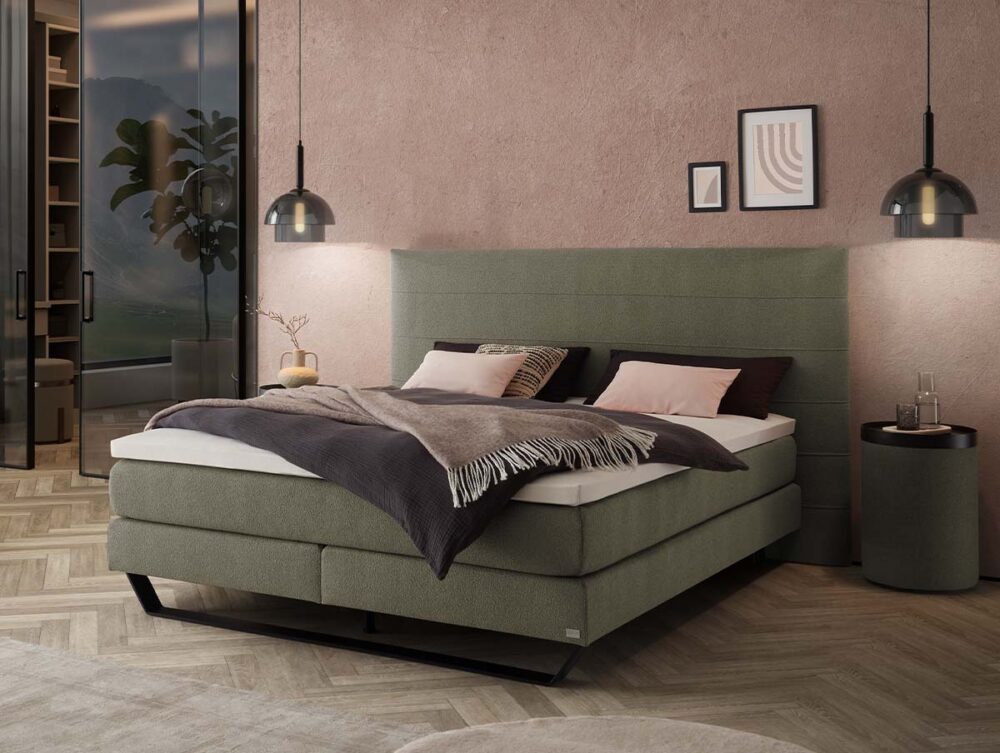 Modernes Schlafzimmer mit einem grün gepolsterten Bett, grauen und beigen Kissen, einer grauen Decke, Hängeleuchten, einem runden Beistelltisch, Wandkunst und einer Glastür, die zu einem begehbaren Kleiderschrank führt, in dem Pflanzen zu sehen sind.