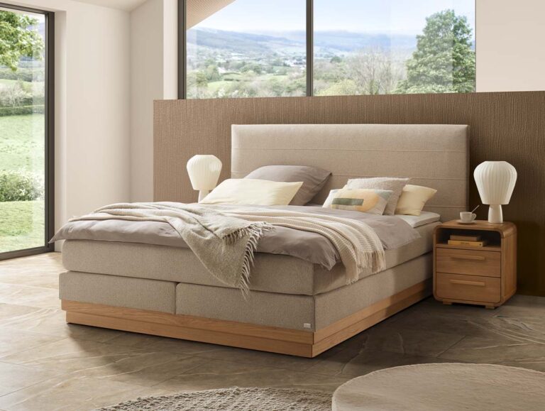Modernes Schlafzimmer mit einem großen beigen Bett, neutraler Bettwäsche, zwei weißen Lampen auf hölzernen Nachttischen und großen Fenstern, die eine grüne Landschaft nach draußen zeigen. Das Zimmer hat einen ruhigen, minimalistischen Stil mit natürlichem Licht.