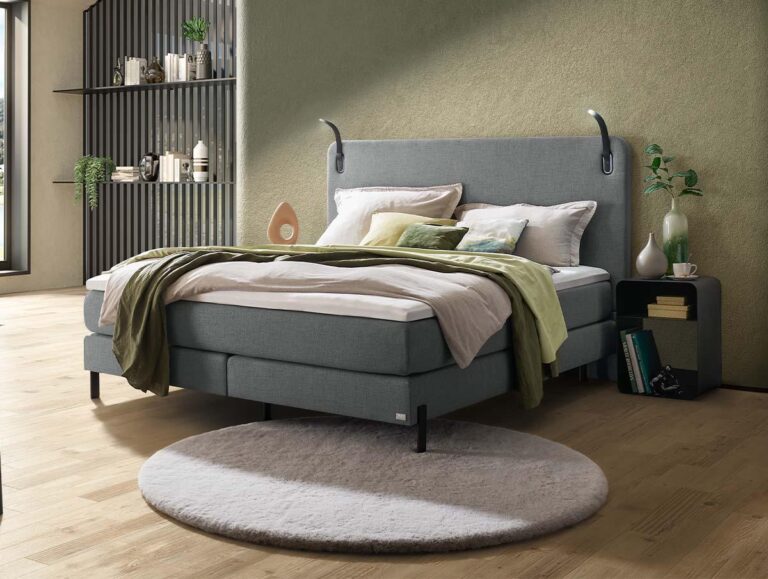 Ein modernes Schlafzimmer mit einem grauen Polsterbett, grüner und weißer Bettwäsche, einem runden beigen Teppich und einem Nachttisch. Auf der linken Seite befinden sich Regale mit Büchern und Dekorationsartikeln. Der Raum hat einen Holzboden und weiches natürliches Licht.