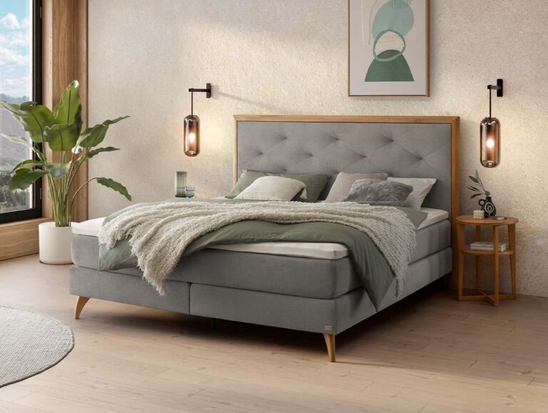 Ein modernes Schlafzimmer mit einem grauen Polsterbett, grüner und beiger Bettwäsche, Nachttischen, Hängelampen, einer Topfpflanze und abstrakter Wandkunst. Natürliches Licht fällt durch ein großes Fenster ein.