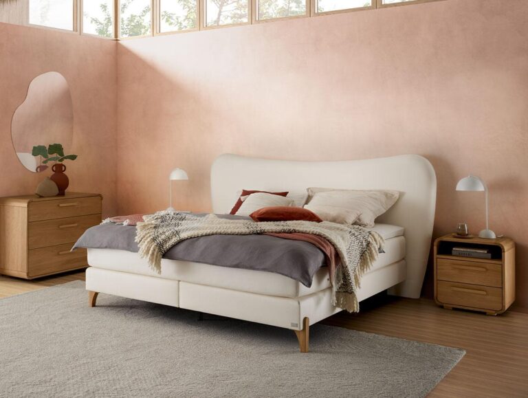 Ein gemütliches Schlafzimmer mit einem cremefarbenen Bett, mehrlagigen Decken, neutralen Kissen, hölzernen Nachttischen, Tischlampen, einer Kommode mit einer Vase und einer Pflanze, einem großen Teppich und pastellrosa Wänden mit hohen Fenstern, die natürliches Licht hereinlassen.