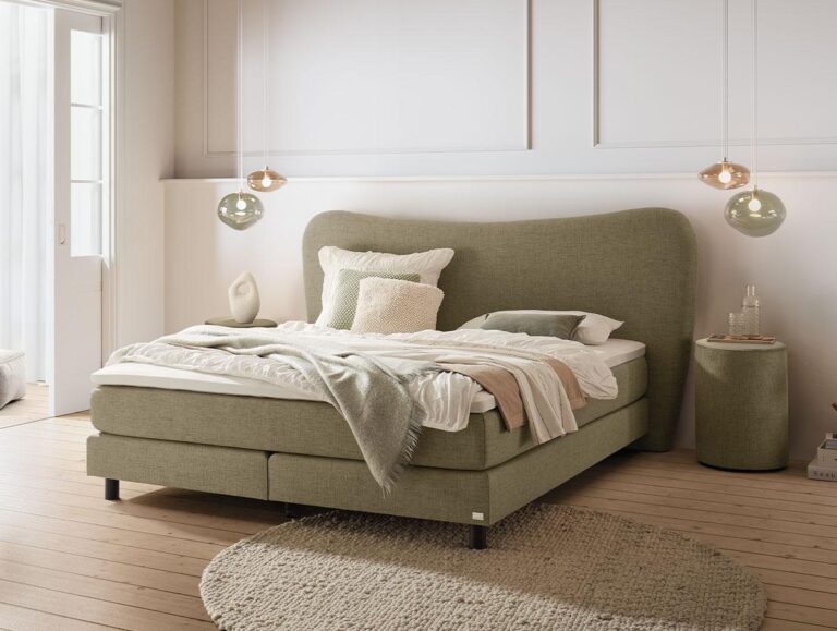 Ein modernes Schlafzimmer mit einem grün gepolsterten Bett mit mehreren Decken und Kissen, einem runden Nachttisch, zwei hängenden Pendelleuchten aus Glas, Holzböden und sanftem Tageslicht aus einem nahe gelegenen Fenster.