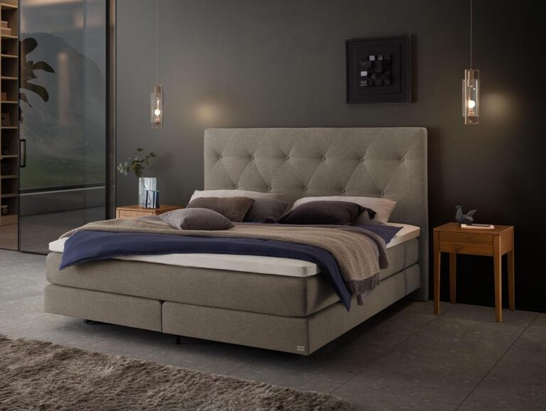 Ein modernes Schlafzimmer mit einem grauen Polsterbett, Lagenbettwäsche, zwei hölzernen Nachttischen mit Pendelleuchten darüber und einer dunklen Akzentwand. Auf dem Boden liegt ein Teppich, und auf der linken Seite ist ein Regal zu sehen.