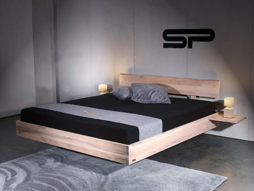 Ein modernes, minimalistisches Holzpritschenbett mit einem niedrigen Kopfteil, schwebenden Nachttischen und zwei kleinen Lampen. Das Bett ist mit grauer und schwarzer Bettwäsche und zwei grauen Kissen ausgestattet und steht in einem schlichten Raum mit Betonwänden und einem grauen Teppichboden.