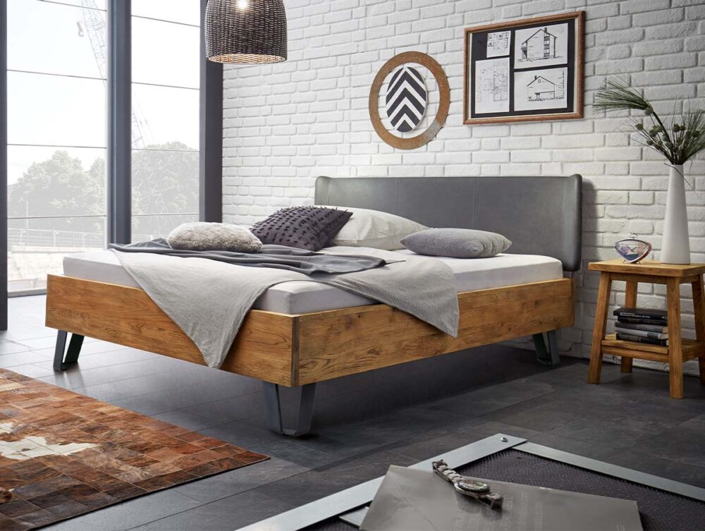 Ein modernes Schlafzimmer mit einem Bettgestell aus Holz, einem grauen Kopfteil und neutraler Bettwäsche. Das Zimmer hat weiße Backsteinwände, große Fenster, Wandkunst, einen hölzernen Beistelltisch mit Dekoration und eine hängende geflochtene Lampe.