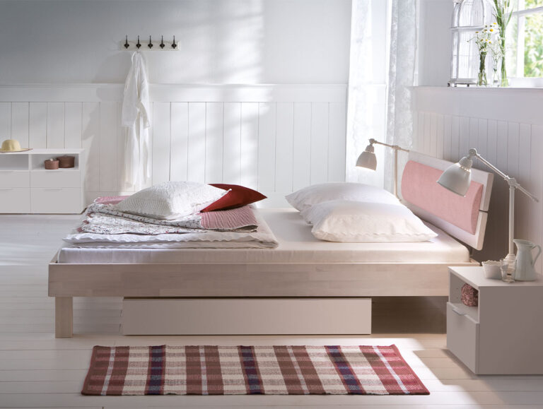 Ein helles, minimalistisches Schlafzimmer mit dem Hasena Bett "Wood-Line Classic Cantu 3", weißen Wänden und Möbeln, knackig weißer Bettwäsche mit einem roten Akzentkissen, Wandhaken mit einem Bademantel und Sonnenlicht, das durch das Fenster fällt.