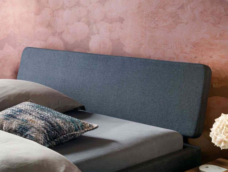Ein modernes Bett mit einem dunkelgrau gepolsterten Kopfteil, grauer Bettwäsche, einem gemusterten Akzentkissen und einem Nachttisch mit einem Strauß weißer Blumen; die Wand dahinter ist mit einer zartrosa Blumentapete versehen.