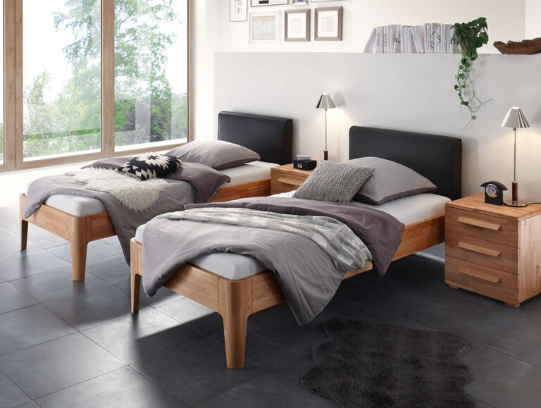 Ein modernes Schlafzimmer mit zwei Einzelbetten, Holzmöbeln, grauer Bettwäsche, großen Fenstern mit Blick auf den Garten, einem Schreibtisch, Lampen und gerahmten Bildern an einer weißen Wand. Der Boden ist dunkel gefliest und mit zwei Teppichen ausgelegt.