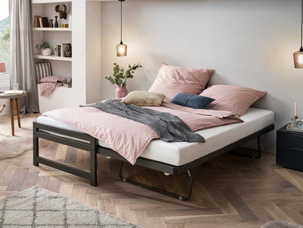 Ein modernes Schlafzimmer mit einem ordentlich gemachten Bett mit rosa und grauer Bettwäsche, zwei Pendelleuchten, einem kleinen schwarzen Nachttisch und offenen Regalen mit Dekoration und Pflanzen. Das Licht aus einem Fenster erhellt den gemütlichen Raum.