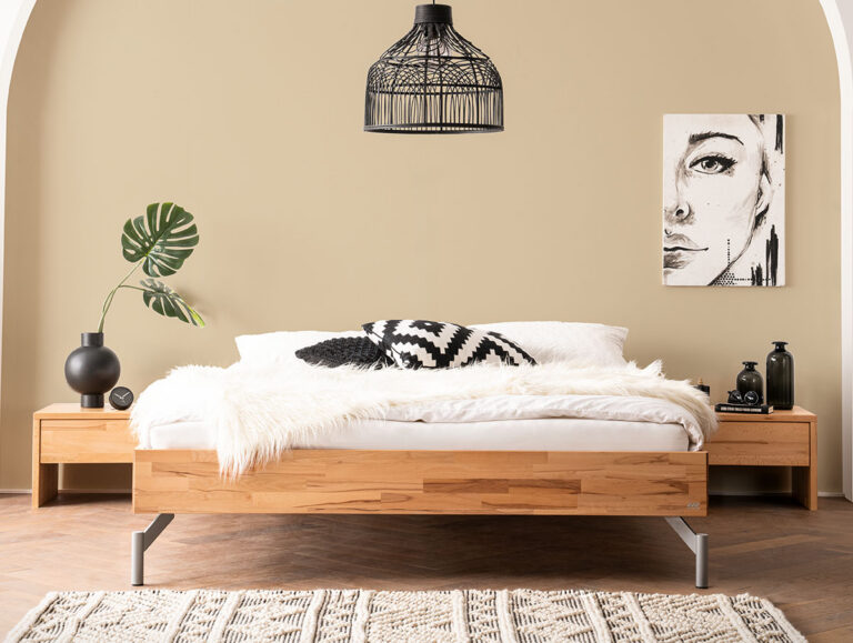 Minimalistisches Schlafzimmer mit einem Holzbett, weißer Bettwäsche, schwarz-weißen Kissen, einer Pelzdecke, passenden Nachttischen aus Holz, einer schwarzen Pendelleuchte und einem modernen Kunstporträt an der beigen Wand. Ein geometrischer Teppich bedeckt den Boden.