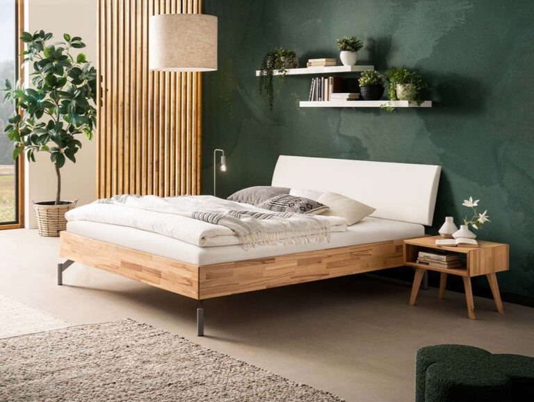 Ein modernes Schlafzimmer mit einem hölzernen Bettgestell, weißer Bettwäsche, einem hölzernen Nachttisch mit einer Lampe, einer grünen Akzentwand, Regalen mit Dekoration, einer großen Topfpflanze und natürlichem Licht aus einem Fenster.