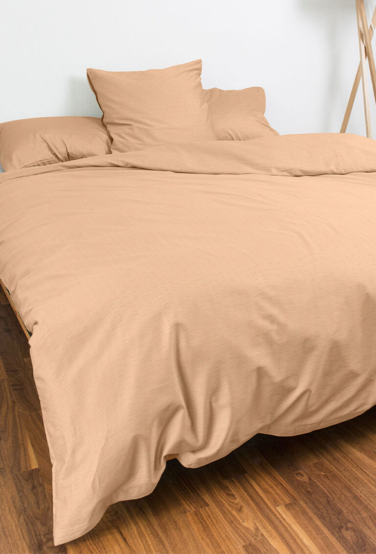 Ein ordentlich gemachtes Bett mit beigem Bettzeug und zwei Kissen, auf einem Holzboden in einem minimalistischen Zimmer mit weißen Wänden.