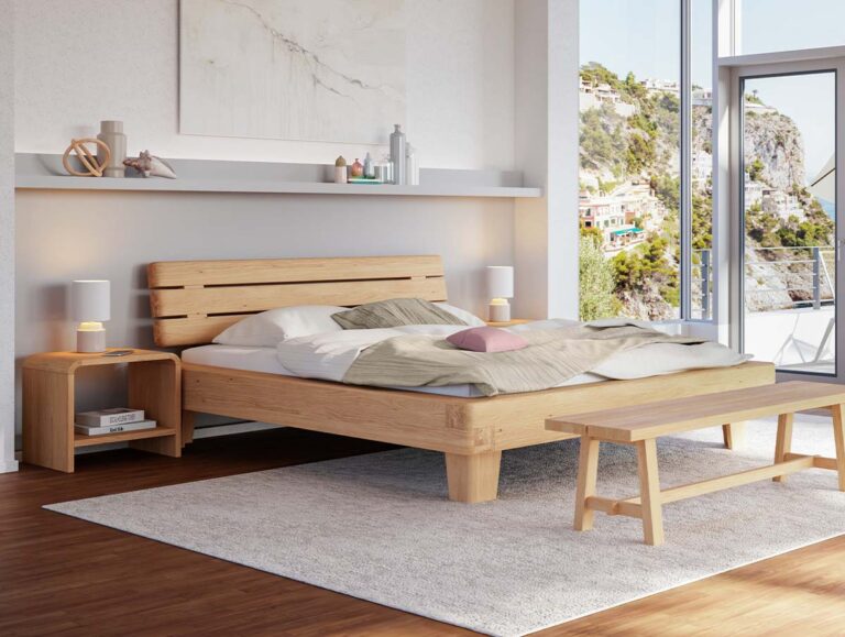 Modernes Schlafzimmer mit einem Bettgestell aus Holz, zwei Nachttischen mit Lampen, einer Bank und großen Fenstern, die einen malerischen Blick auf die Hügel bieten. Neutrale Farbtöne und eine schlichte Einrichtung schaffen eine gemütliche, einladende Atmosphäre.