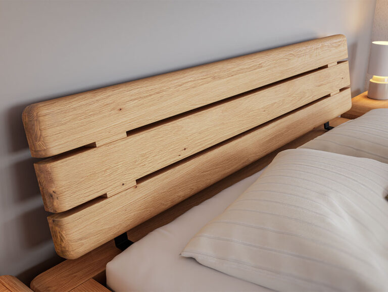 Eine Nahaufnahme eines Bettkopfendes aus Holz mit einer glatten, hellen Holzoberfläche und horizontalen Latten, daneben ein weißes Kissen und eine kleine Nachttischlampe.