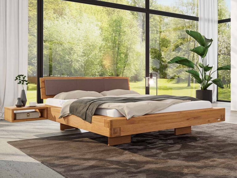 In einem hellen Zimmer mit großen Fenstern, grünen Pflanzen und einem dunkelbraunen Teppich steht ein modernes Holzbett mit beiger Bettwäsche. Das Sonnenlicht fällt durch die Bäume draußen herein.