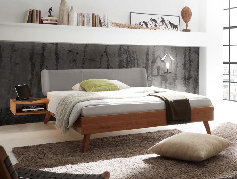 Modernes Schlafzimmer mit einem Bettgestell aus Holz, einem grauen Kopfteil, neutraler Bettwäsche, einem schwebenden Nachttisch, Wandbildern, Büchern, einem gemütlichen Teppich, einem Kissen auf dem Boden und großen Fenstern, durch die Tageslicht hereinströmt.