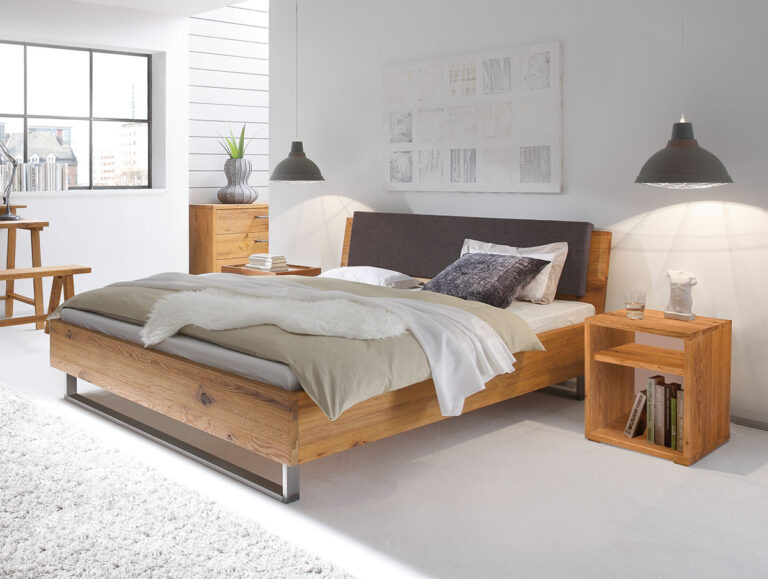 Modernes Schlafzimmer mit einem Holzbett, einem grauen Kopfteil, beiger Bettwäsche, zwei Pendelleuchten, einer flauschigen Decke, einem Nachttisch mit Deko und einem großen Fenster, durch das Tageslicht hereinströmt. Weiße Wände und ein Teppichboden sorgen für eine helle, luftige Atmosphäre.