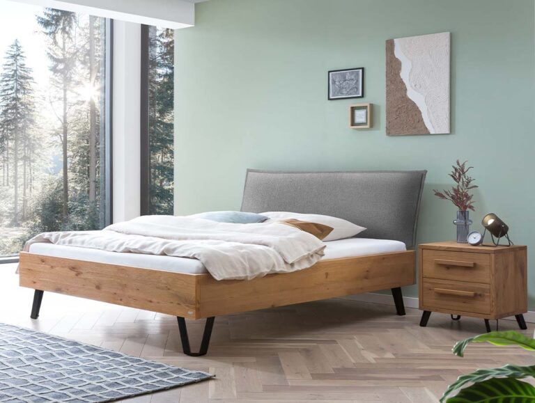 Ein modernes Schlafzimmer mit einem Holzbett, einem grauen Kopfteil, weißer Bettwäsche und einem Nachttisch aus Holz. Auf dem Nachttisch stehen eine Lampe, Bücher und Dekoartikel. Durch die großen Fenster sieht man Bäume draußen. An der hellgrünen Wand hängt ein Bild.