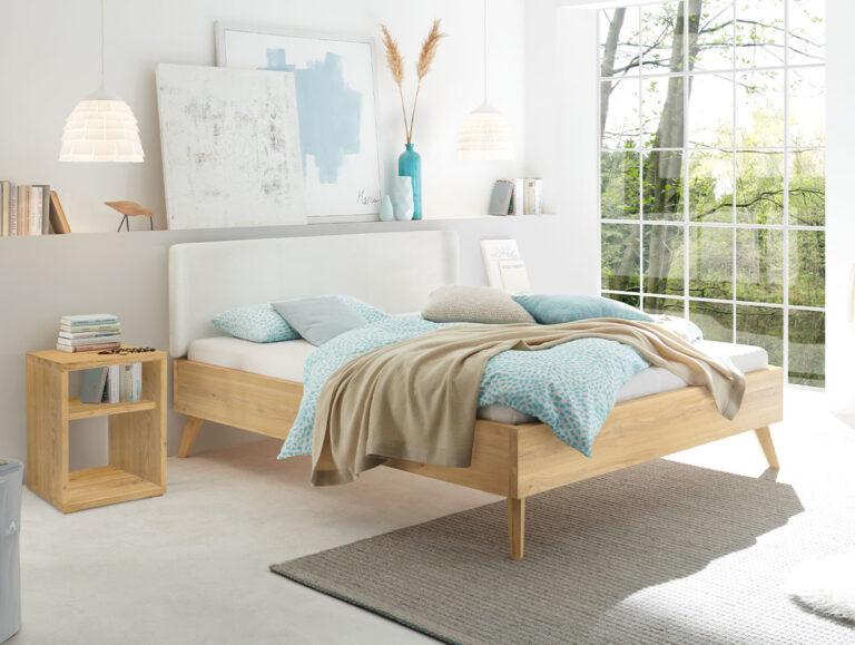 Ein modernes Schlafzimmer mit einem Holzbett, blauer und beiger Bettwäsche, einem Nachttisch, abstrakter Kunst, Pendelleuchten, Büchern und großen Fenstern mit Blick ins Grüne.