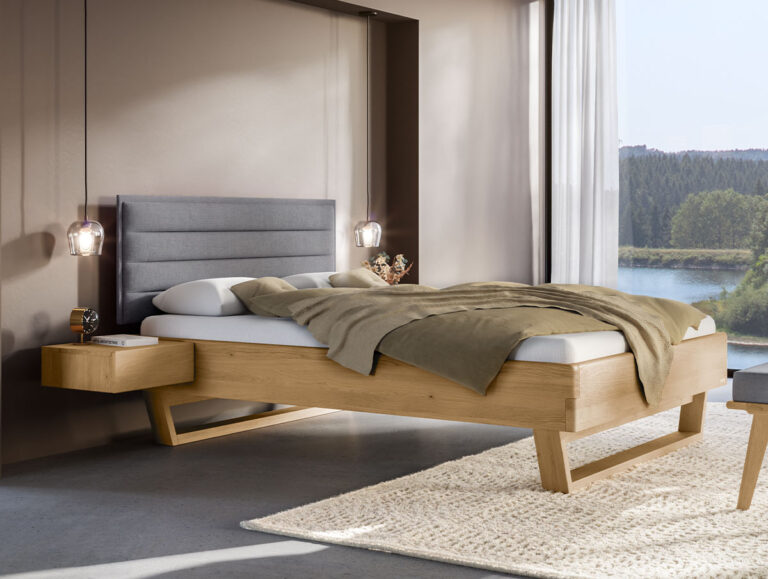 Modernes Schlafzimmer mit einem Hasena-Massivholzbett „Eiche-Bianco“ 9.104 (Kopie), einem grauen gepolsterten Kopfteil, beiger Bettwäsche, einem schwebenden Nachttisch, hängenden Pendelleuchten, einem großen Fenster und einem cremefarbenen Teppich auf einem Betonboden.