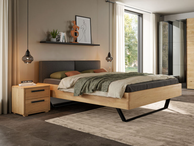 Ein modernes Schlafzimmer mit dem Hasena-Massivholzbett „Eiche-Bianco“ 9.132 – einem stilvollen, schwebenden Holzbett, kombiniert mit einem dunklen Kopfteil, einem Nachttisch mit Schubladen, Pendelleuchten, einem Deko-Regal, großen Fenstern und einem beigen Teppich, der elegant auf dem dunklen Boden platziert ist.