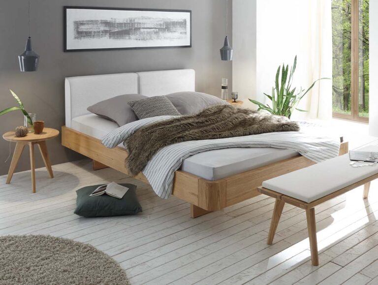 Ein modernes Schlafzimmer mit dem eleganten Hasena-Massivholzbett „Eiche-Bianco“ (Art.-Nr. 9.122), ergänzt durch strahlend weiße Bettwäsche, graue und braune Decken, üppige Pflanzen, einen runden Beistelltisch aus Holz, eine Bank und ein großes Fenster, das den Raum mit Tageslicht durchflutet.