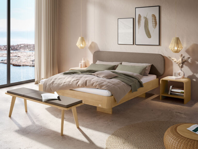 Ein modernes, lichtdurchflutetes Schlafzimmer mit einem großen Bett, grüner Bettwäsche, Holzmöbeln, einer Bank, zwei Hängeleuchten, gerahmten Botanikkunstwerken und einem Fenster mit Blick auf eine felsige Küstenlandschaft.
