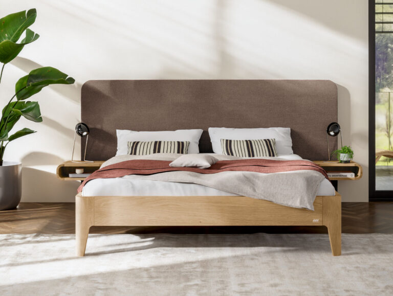 Ein modernes Bett mit Holzrahmen, gepolstertem braunem Kopfteil, gestreiften Kissen und einer roten Tagesdecke. Auf den eingebauten Nachttischablagen stehen zwei kleine Lampen, links davon eine große grüne Pflanze und rechts ein Fenster.