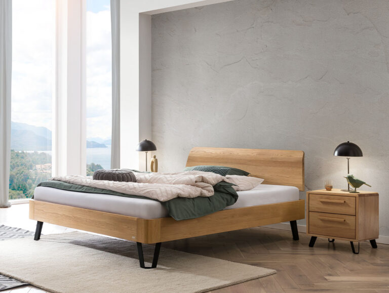 Ein modernes Schlafzimmer mit einem Holzbett, unordentlicher Bettwäsche, einem Nachttisch aus Holz, zwei schwarzen Lampen, großen Fenstern mit Blick auf die Berge und einem minimalistischen Einrichtungsstil.