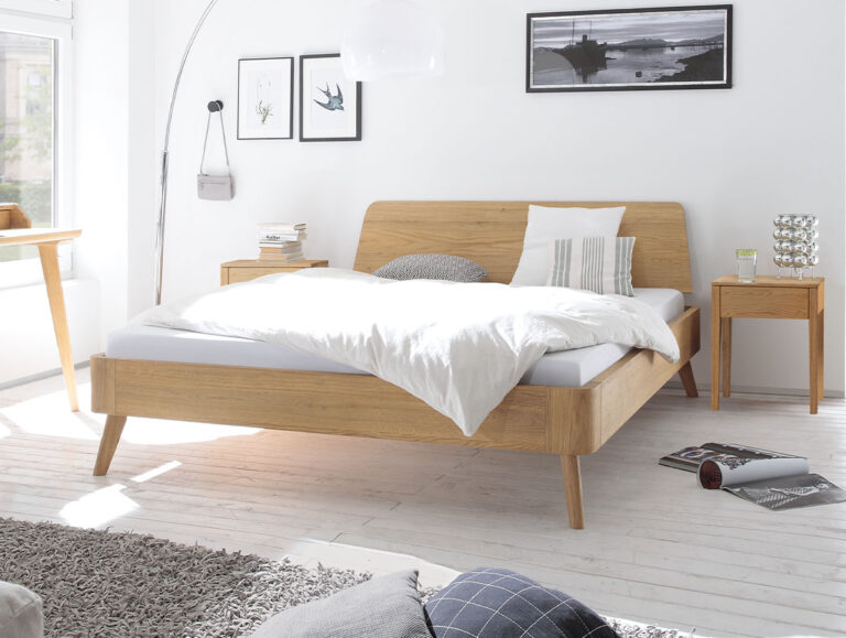 Ein helles, modernes Schlafzimmer mit einem Hasena Massivholzbett „Eiche-Bianco“ (Art.-Nr. 9.112) und frischer, weißer Bettwäsche, ergänzt durch zwei Nachttische aus Holz und gerahmte Kunstwerke an den weißen Wänden. Sonnenlicht strömt durch ein großes Fenster herein und unterstreicht die einladende Atmosphäre.